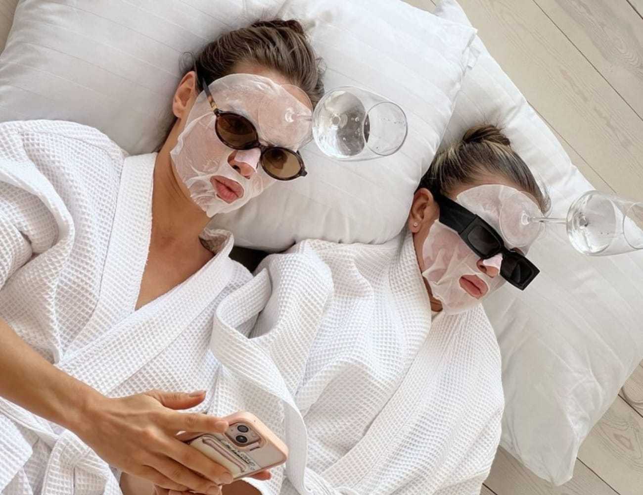 Two people relaxing in robes with face masks and quirky sunglasses, each balancing a glass on their head.